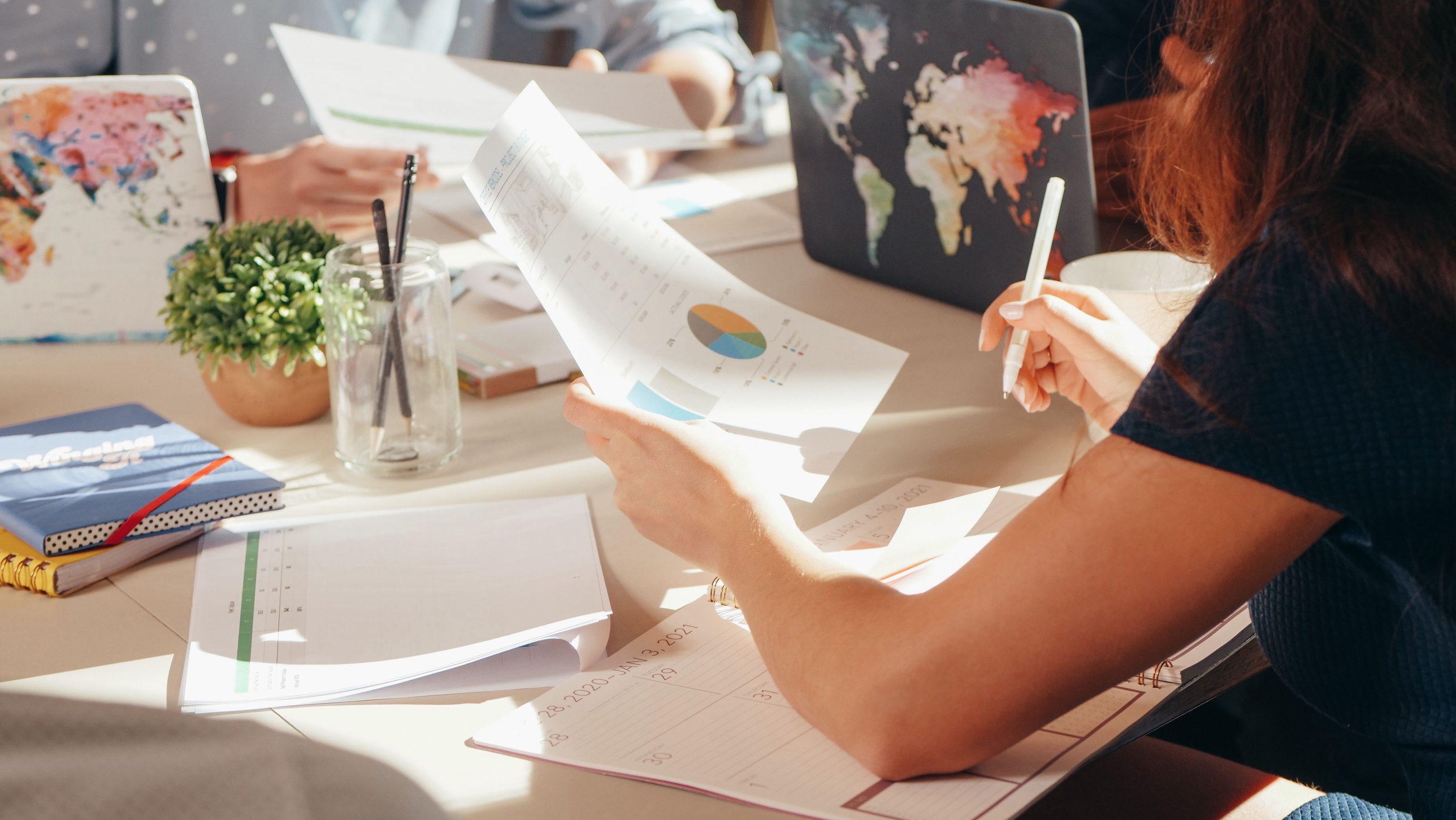 Hands reviewing paperwork and pie charts at a desk with a world map laptop